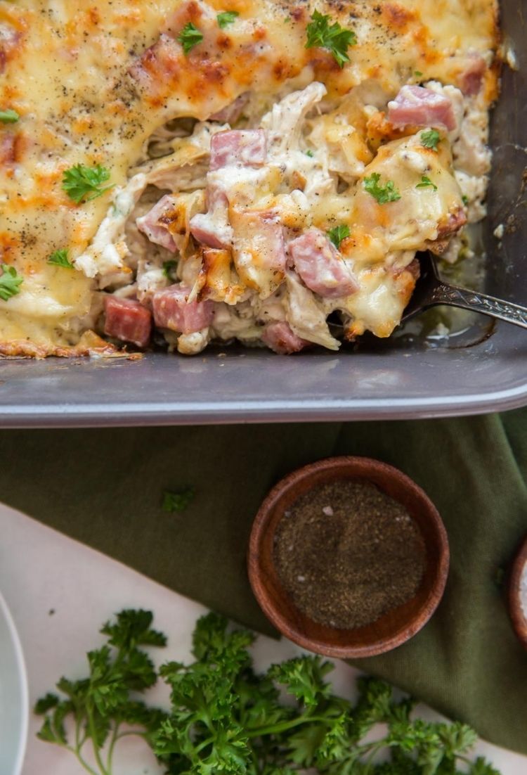 Chicken cordon bleu in a grey baking dish with a silver spoon coming out and all on top of a green towel with seasoning in brown bowl