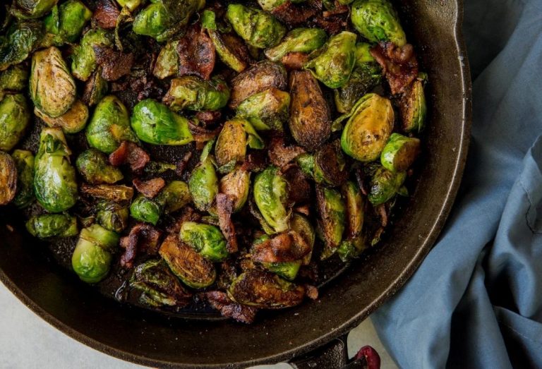 Sauteed Cast Iron Brussels Sprouts with Bacon in a white bowl with red pepper flakes, a spoon, and a fork.