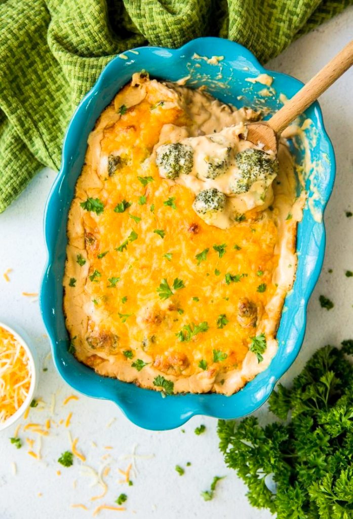 broccoli and cheese casserole in a blue baking dish with a wooden spoon with a green napkin and parsley 