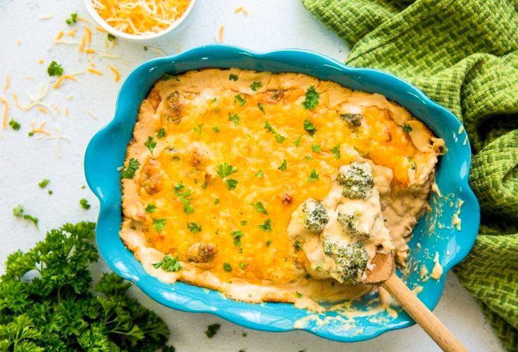 broccoli and cheese casserole in a blue baking dish with a wooden spoon with a green napkin and parsley 