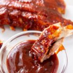 instant pot baby back ribs being dipped into a clear glass filled with BBQ sauce.