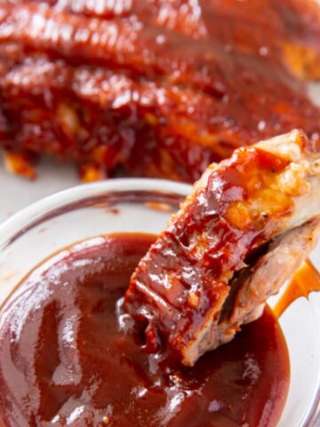 instant pot baby back ribs being dipped into a clear glass filled with BBQ sauce.