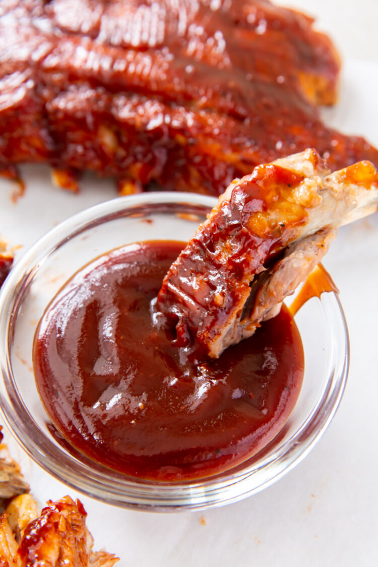 baby back rib being dipped into BBQ sauce in a clear glass container