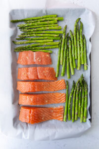 raw salmon and uncooked asparagus on a parchment lined cookie sheet topped with spices