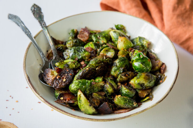 Brussels sprouts with bacon in a white bowl with silverware and a orange napkin