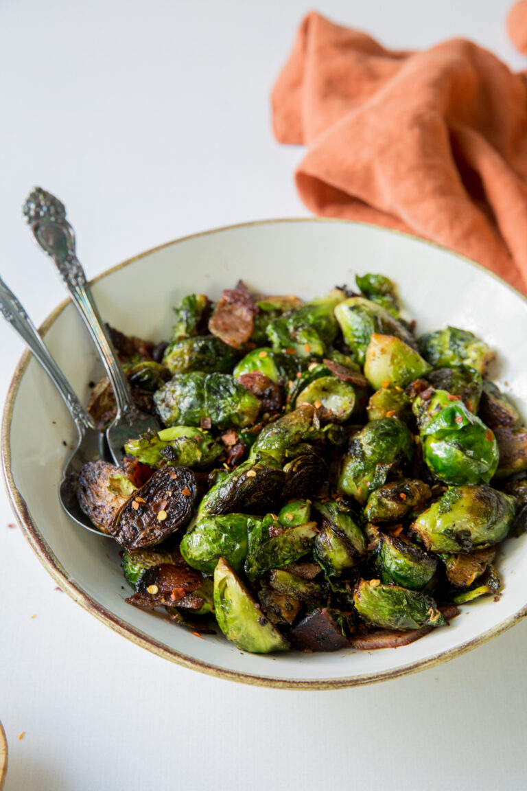 Sauteed Cast Iron Brussels Sprouts with Bacon in a white bowl with red pepper flakes, a spoon, and a fork.