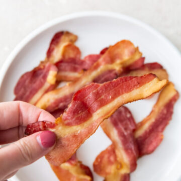 holding a piece of bacon with fingers above a plate of bacon