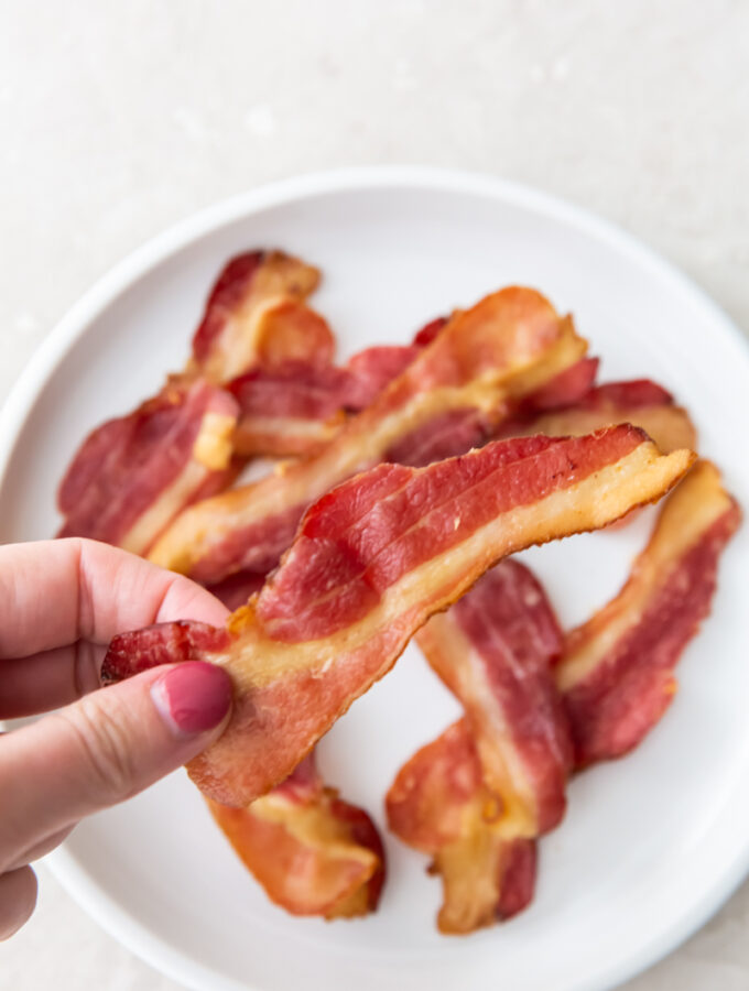 holding a piece of bacon with fingers above a plate of bacon