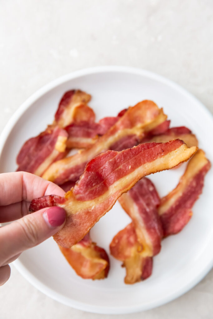 holding a piece of bacon with fingers above a plate of bacon