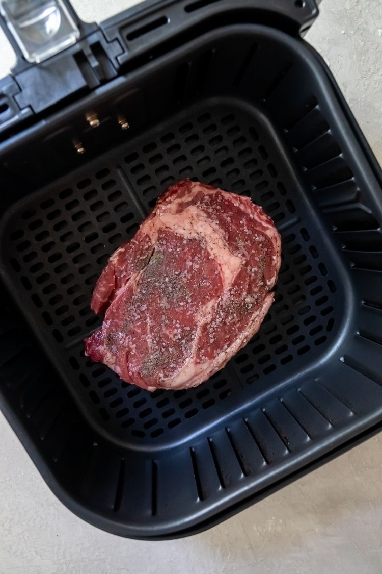 a raw ribeye steak topped with salt and pepper in an air fryer basket
