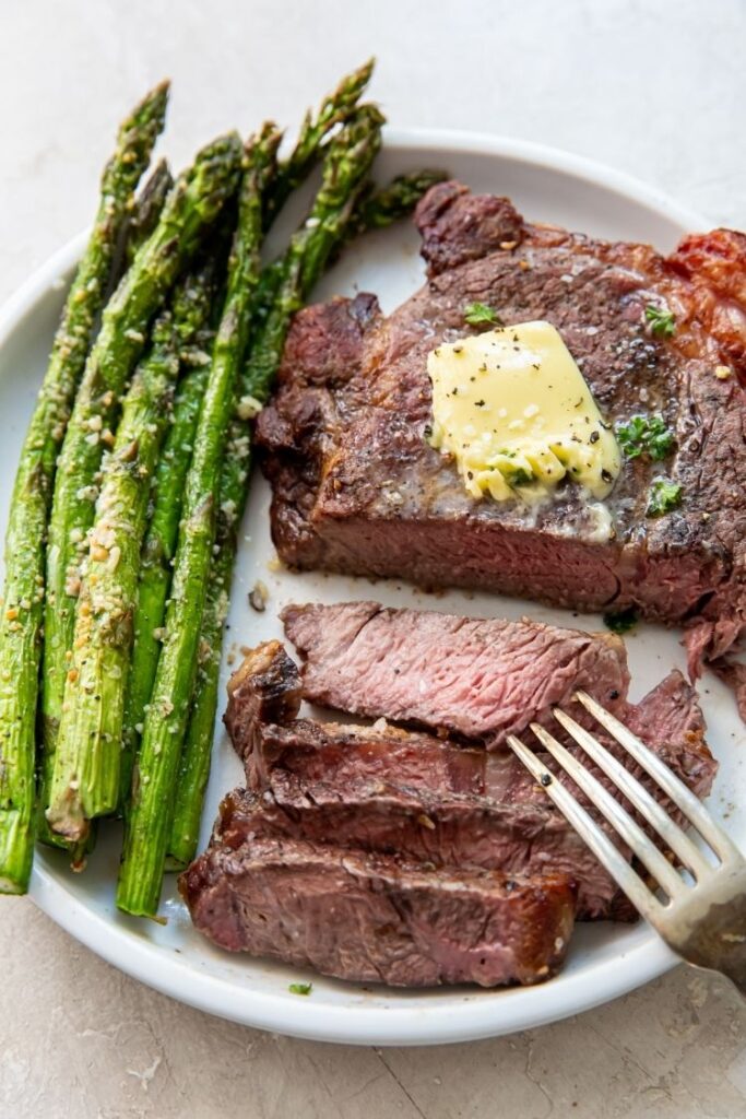 juicy ribeye steak topped with butter served with asparagus and a fork