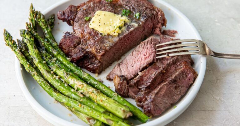 Juicy Air Fryer Ribeye Steak on a white plate with butter, parsley, garlic and asparagus.