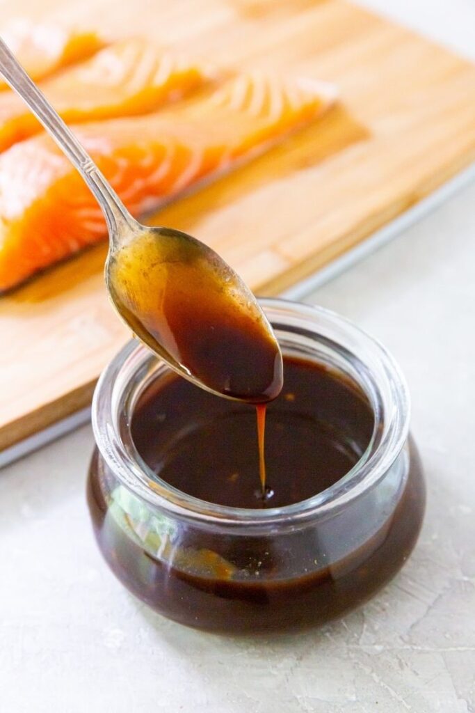 teriyaki sauce in a clear container with a spoon and salmon on a cutting board in background