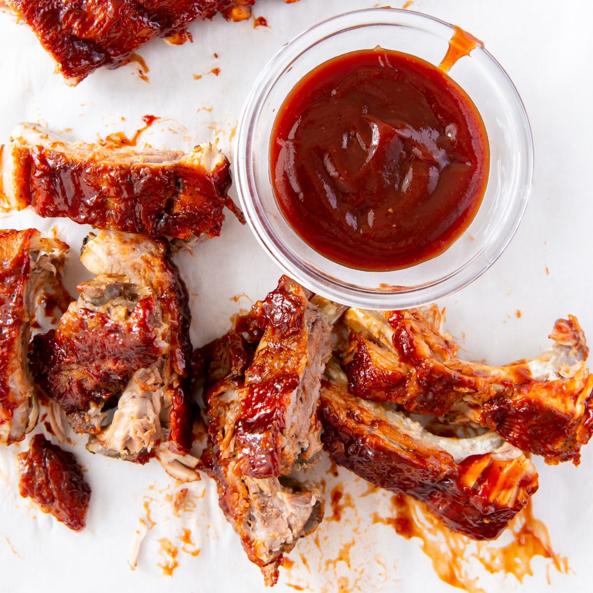 Teriyaki chicken thighs sizzle alongside barbecue ribs on a white surface, with a glass bowl of rich barbecue sauce beside them.