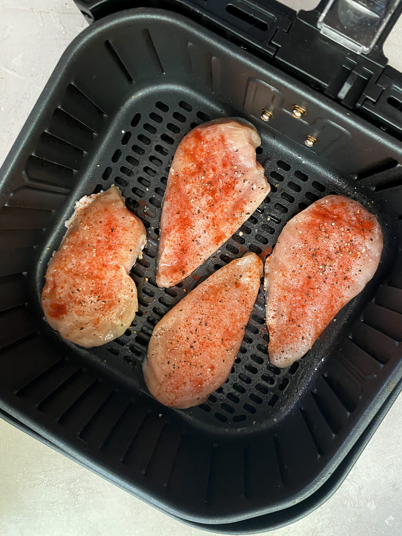 raw 4oz chicken breasts in an air fryer basket with seasonings on top
