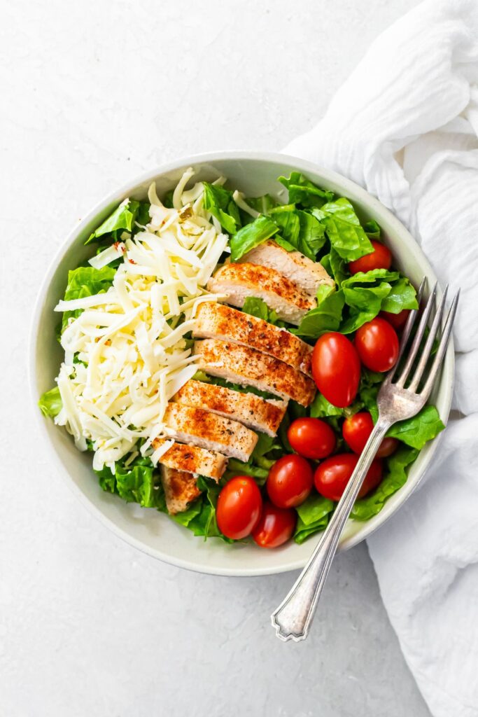 sliced air fried chicken breast on a bed of lettuce with cheese and tomatoes with a fork