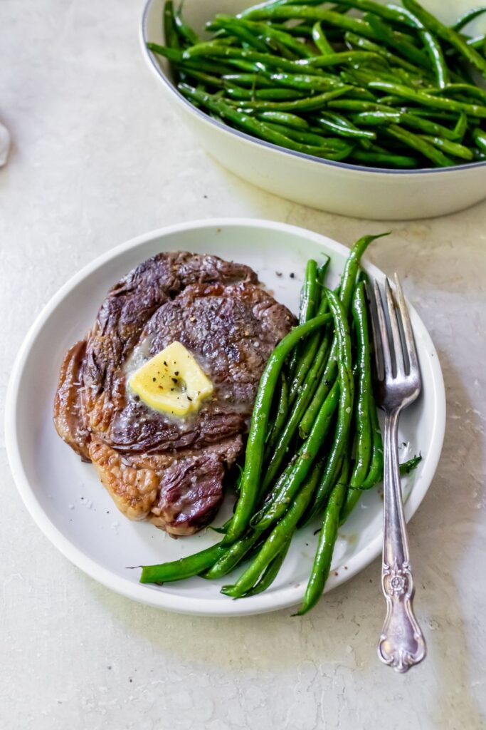 green beans on a white plate with a ribeye and a fork with green beans in the background
