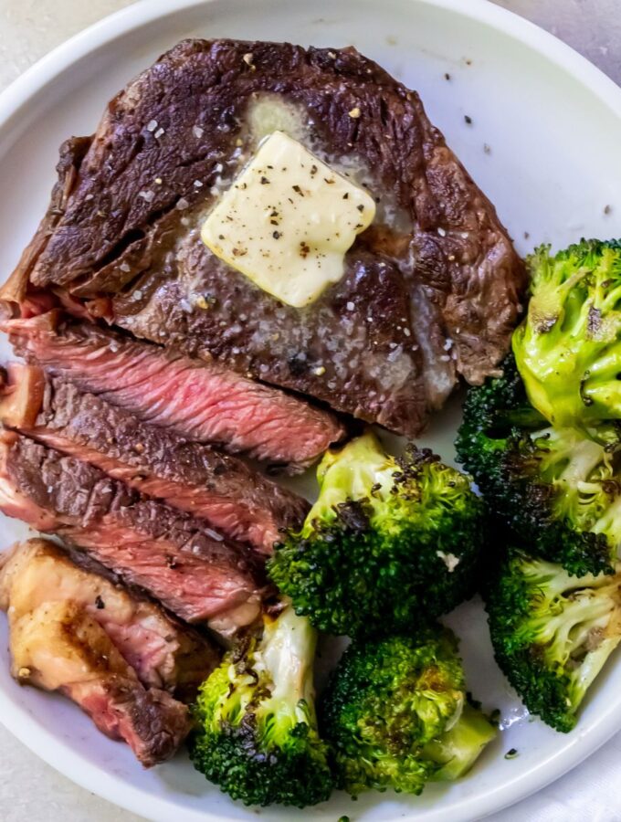 medium rare sliced ribeye steak on a white plate topped with butter and served with broccoli