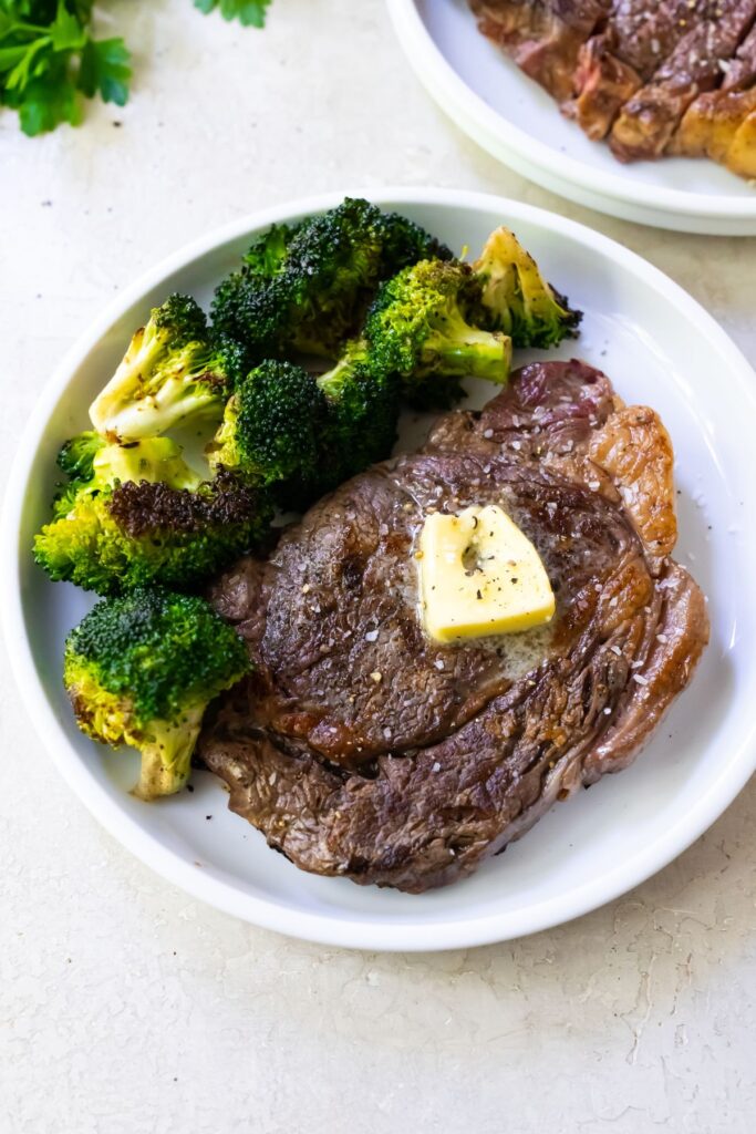 a ribeye steak on a white plate topped with butter and served with a side of broccoli.