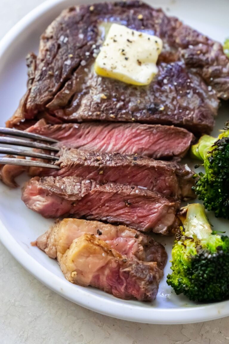 Juicy Ribeye with butter, salt, pepper, and broccoli on a white plate with a fork.