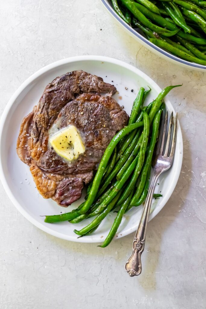 ribeye steak on a white plate with green beans and topped with butter.