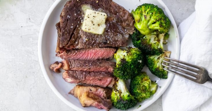 sliced medium rare ribeye on a white plate with broccoli topped with butter