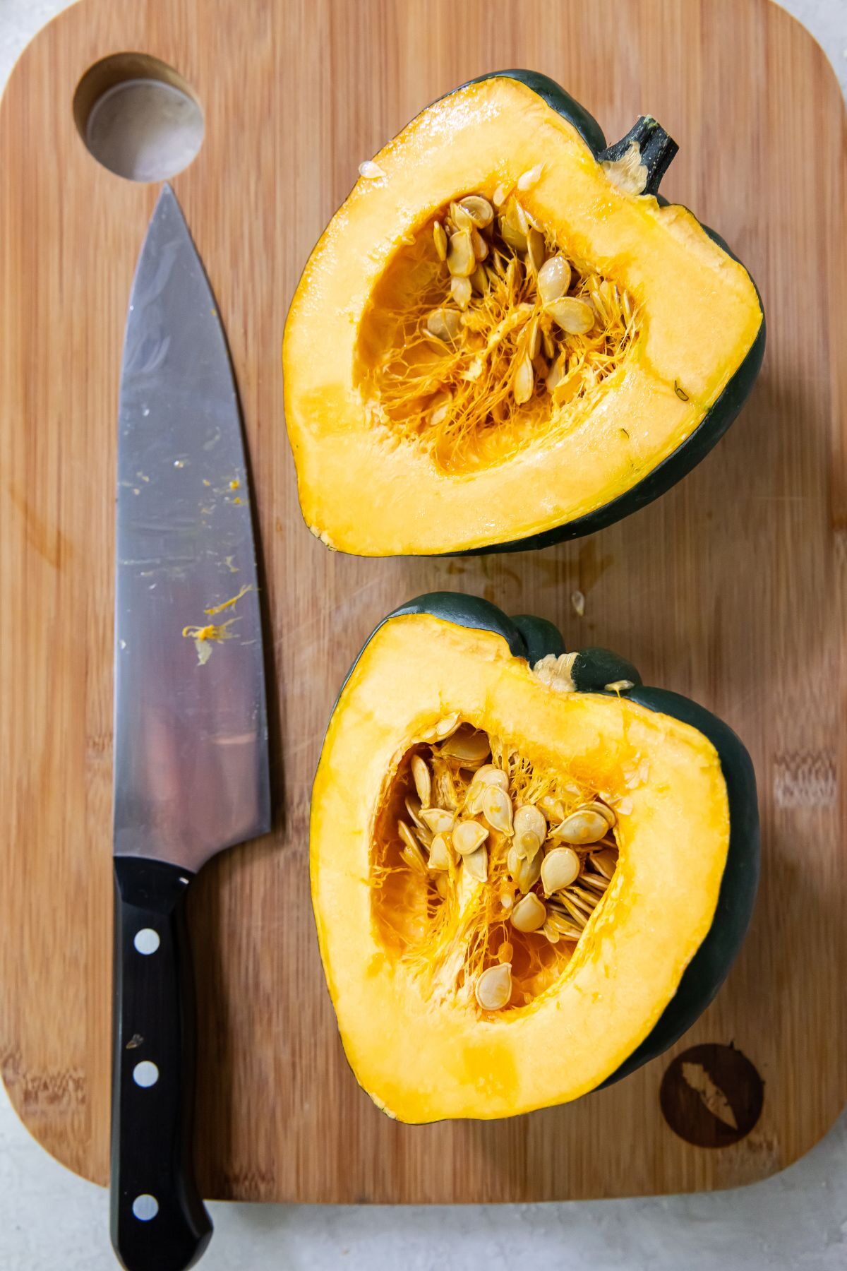 Halved acorn squash with a chef knife to the left both on a wooden cutting board.