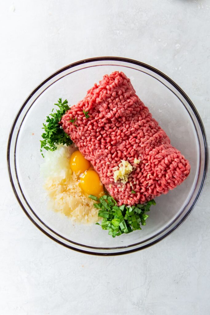 Raw ground beef, egg yolks, garlic, basil and parsley in a glass bowl.