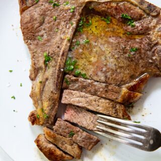 Air Fryer T-bone steak on a white plate butter and parsley on top. fork next to it