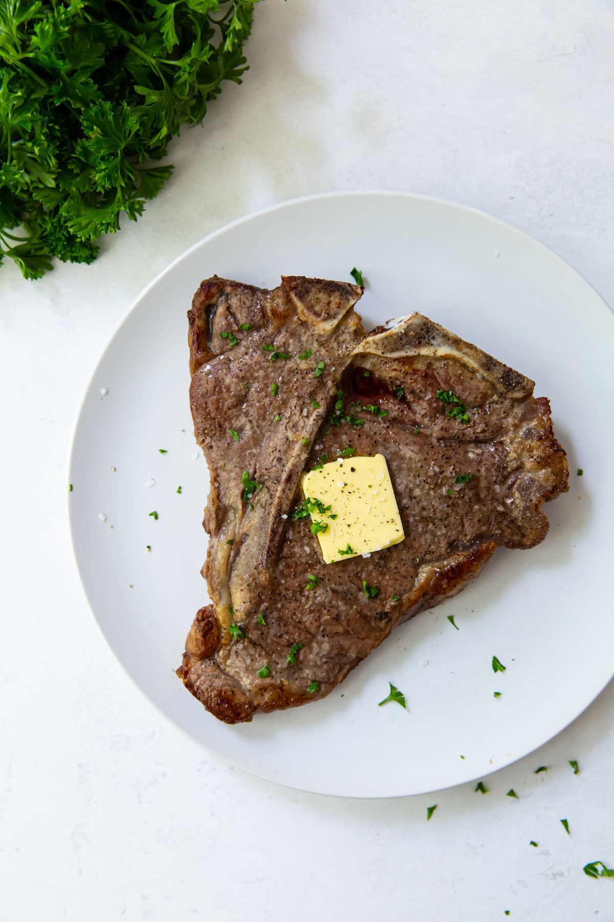 Air Fryer T-bone steak on a white plate butter and parsley on top. parsley in the back