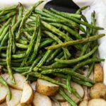 cooking sheet with parchment paper green beans and potatoes one top. spatula holding green beans