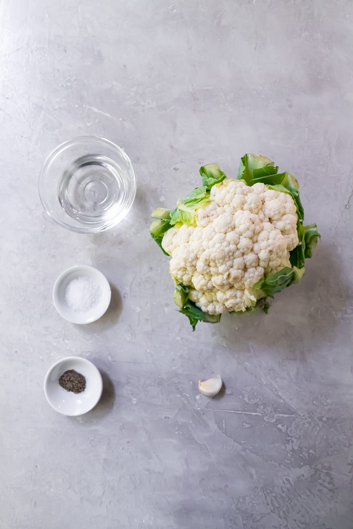 Ingredients consisting of water, salt, pepper, one garlic clove and one head of cauliflower on a white countertop.