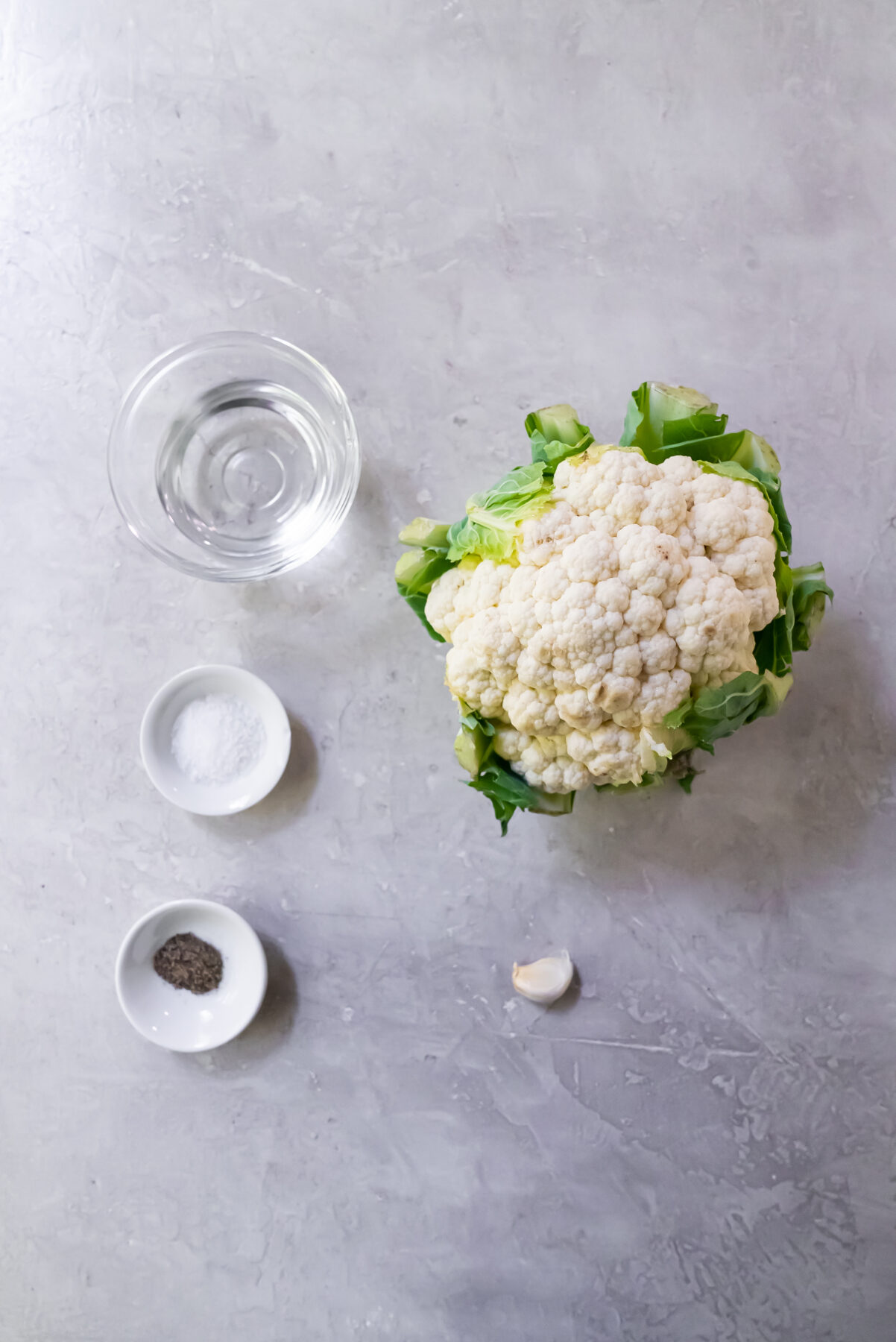 Ingredients consisting of water, salt, pepper, one garlic clove and one head of cauliflower on a white countertop.