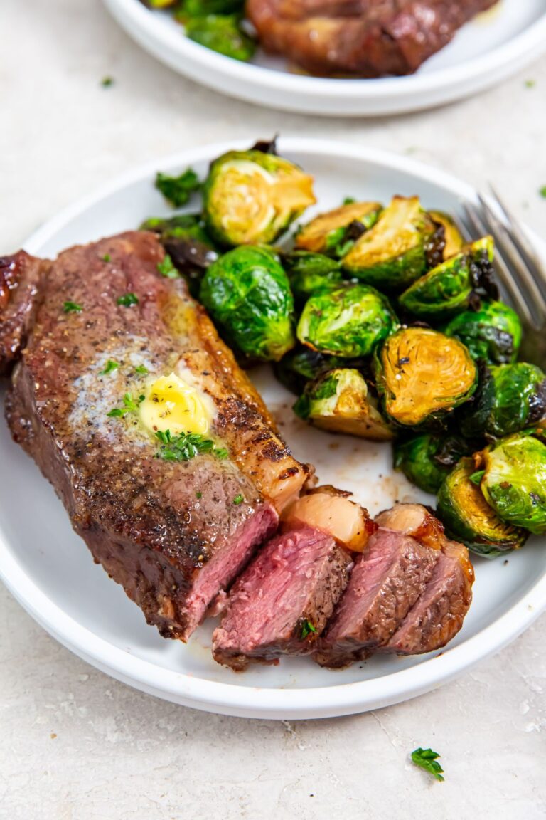 Air Fryer New York Strip Steaks on a white plate with butter, thyme, pepper, salt, brussels sprouts, and a fork.