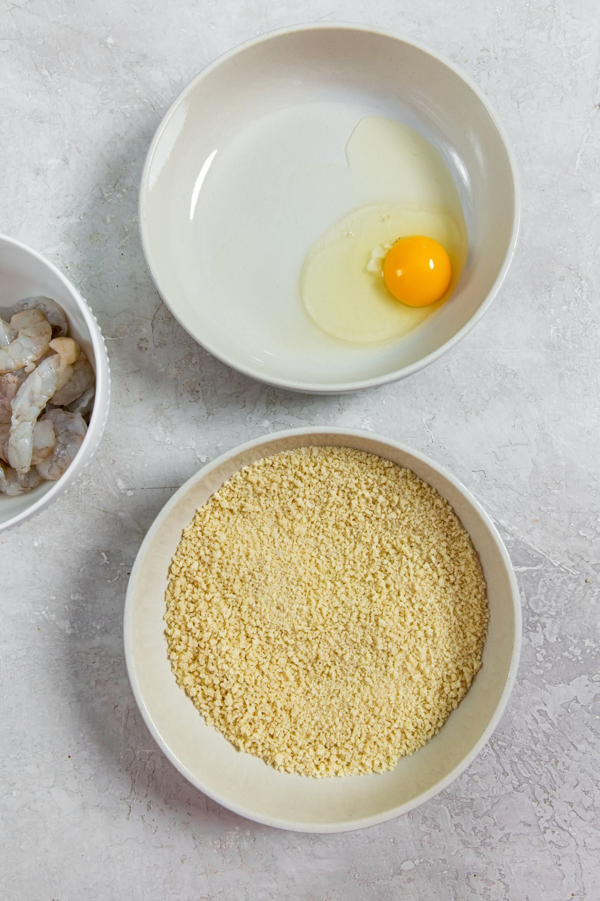 A bowl with an egg and a bowl with panko next to a bowl with raw shrimp