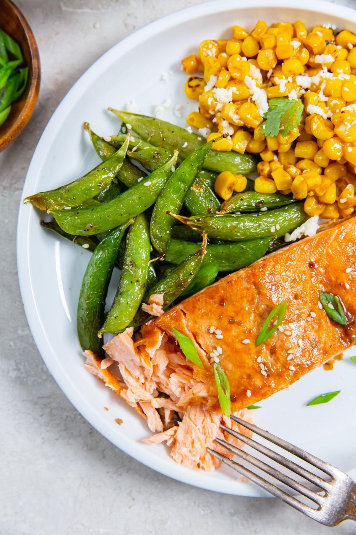 Air Fryer frozen salmon with teriyaki sauce on a white plate with a side of snap peas and a side of corn with a fork in the salmon