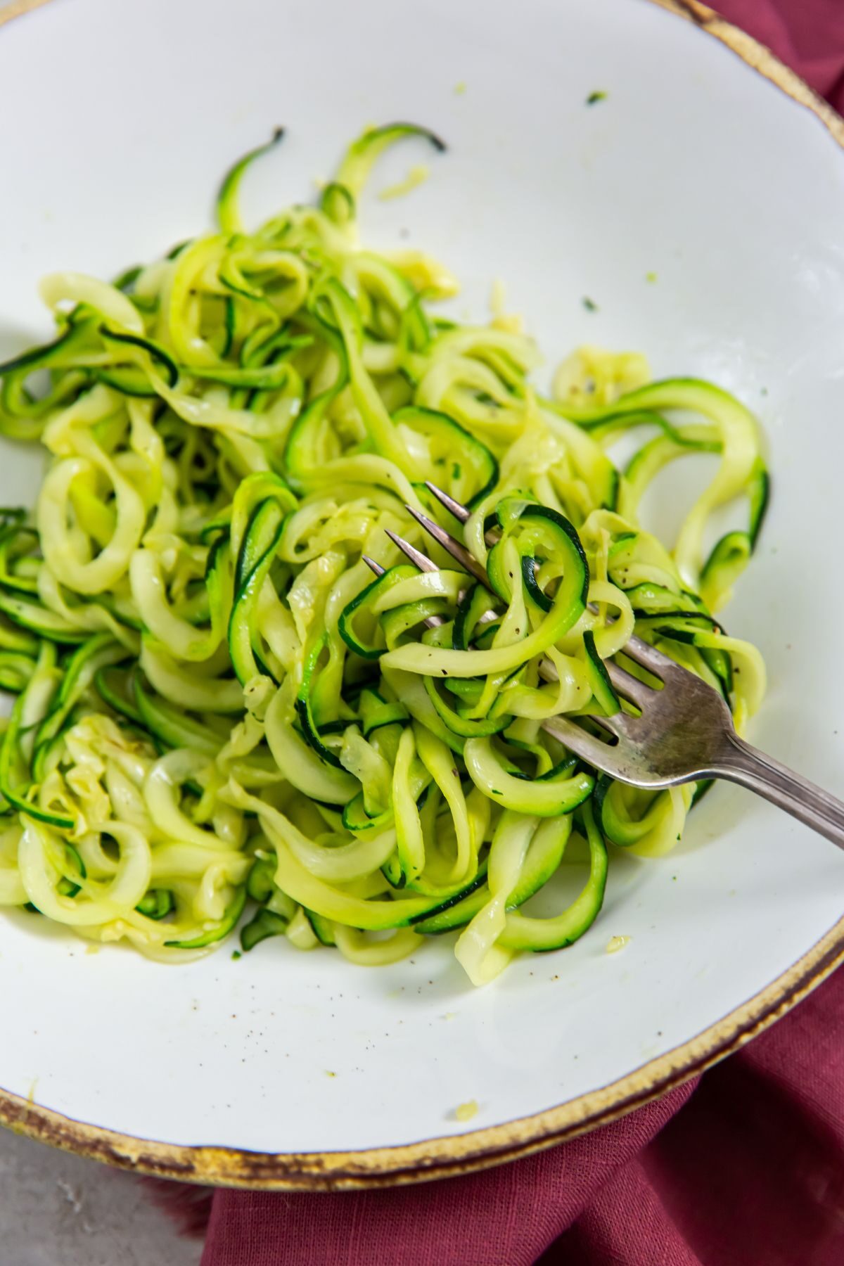 Zoodles in a white bowl with a fork in it with a maroon napkin on the side