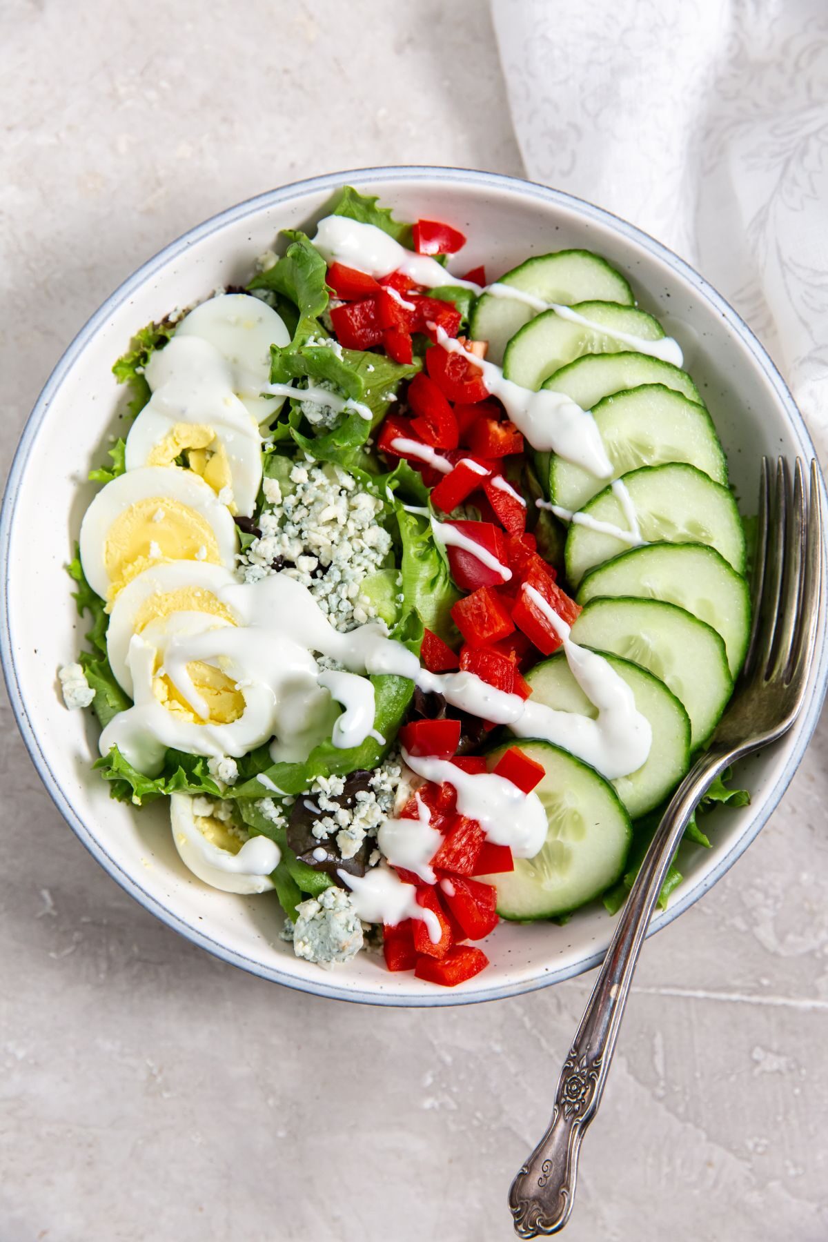 salad in a white bowl with hard boiled eggs, cucumber, red pepper, gorgonzola cheese and ranch dressing.