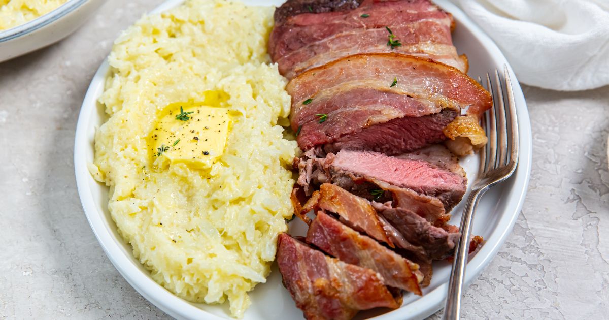 Sliced Air fryer Bacon Wrapped Ribeye Steak with a fork in it and with a side of cheesy mashed cauliflower on a white plate