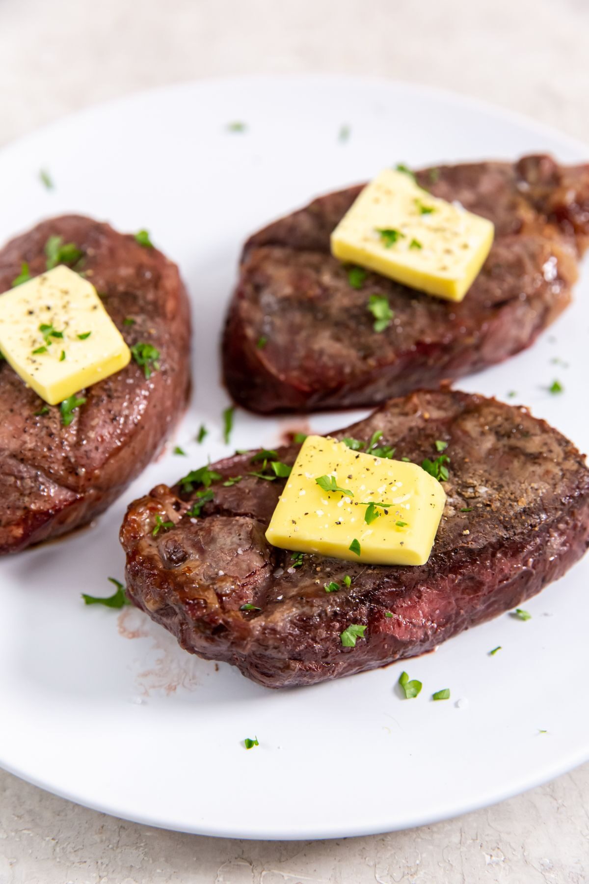Three cooked petite sirloin steaks on a white plate and topped with butter and herbs.