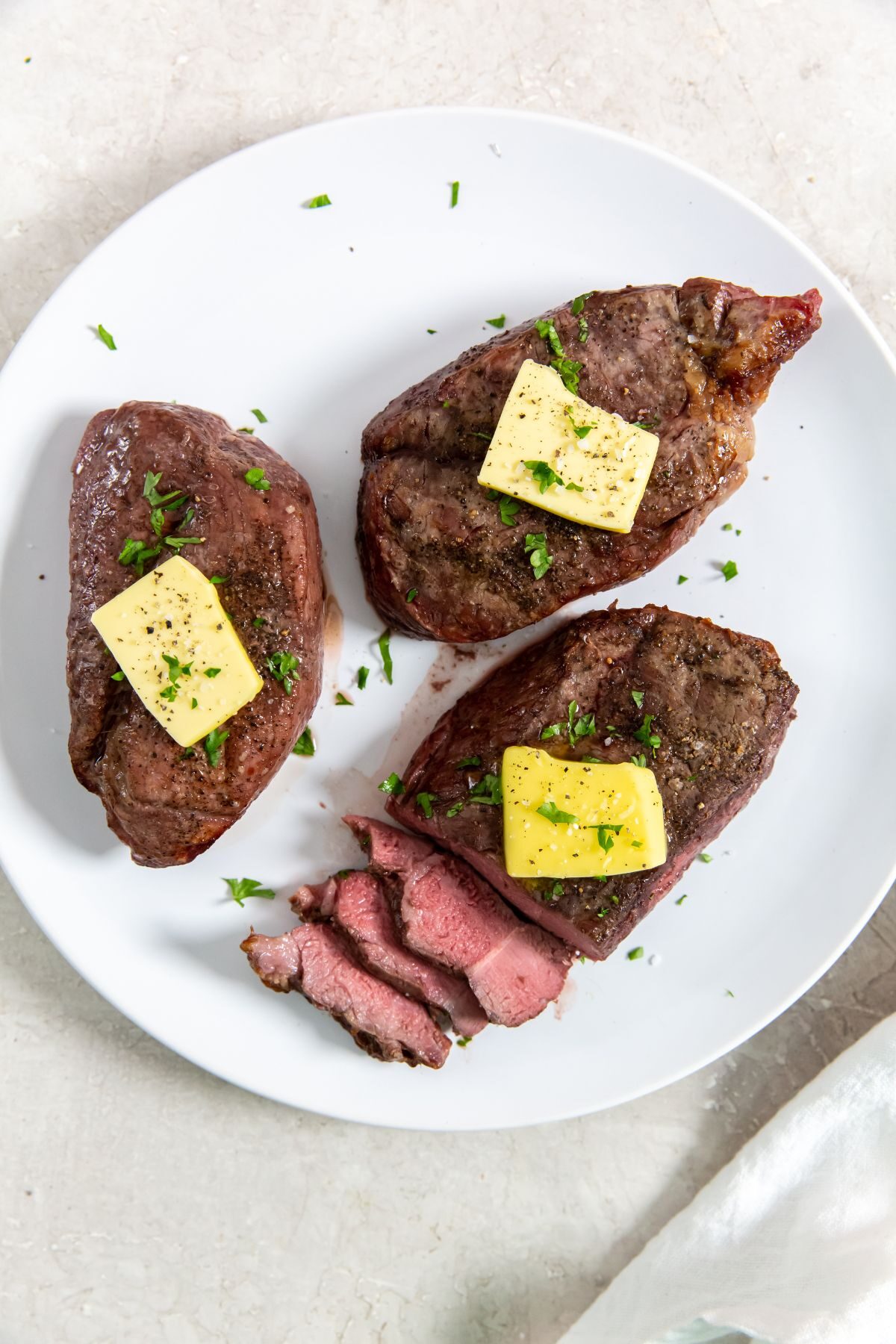 Three cooked and sliced petite sirloin steak topped with butter and herbs on a white plate.