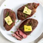Three cooked and sliced petite sirloin steak topped with butter and herbs on a white plate.