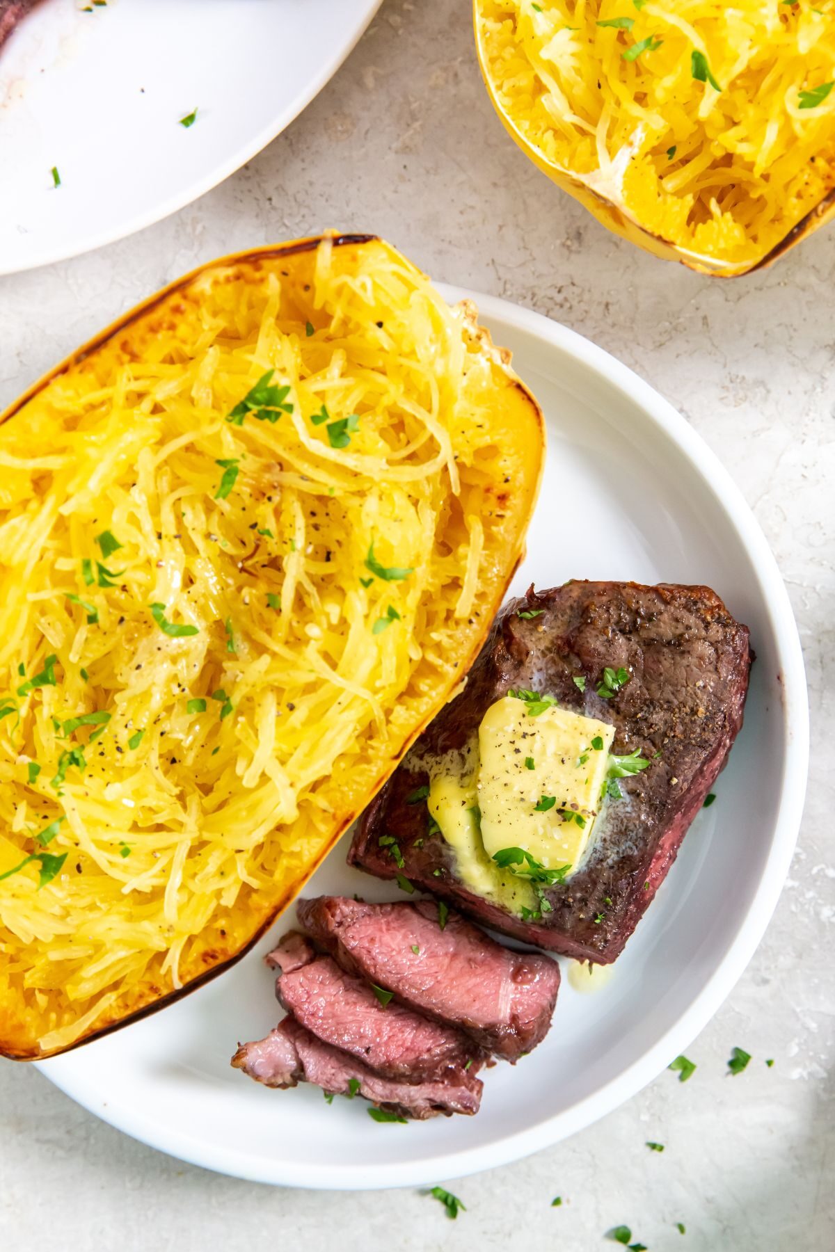 One cooked and sliced petite sirloin steak topped with butter and herbs next to a halved and cooked spaghetti squash on a white plate.