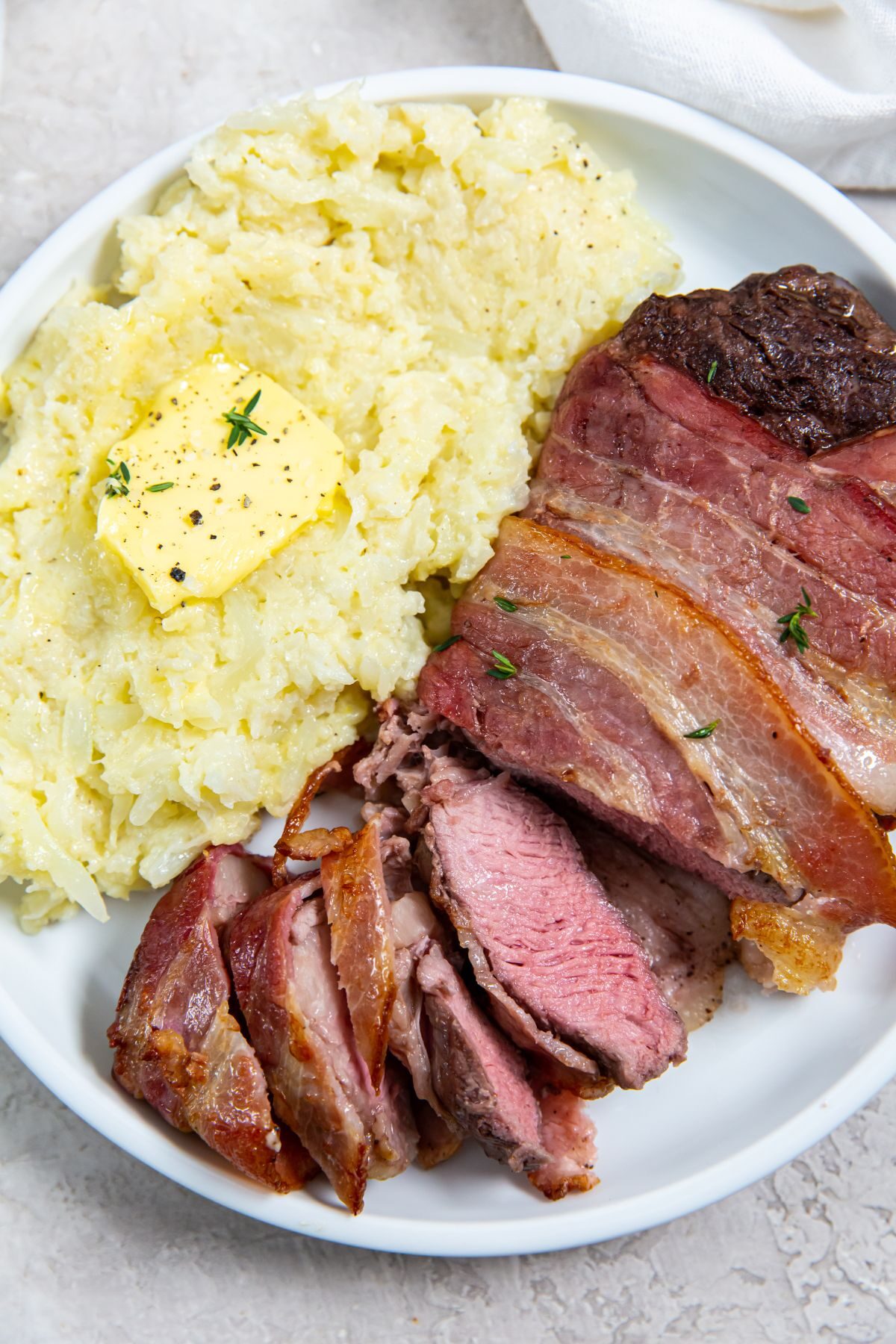 Sliced Air fryer Bacon Wrapped Ribeye Steak with a side of cheesy mashed cauliflower on a white plate