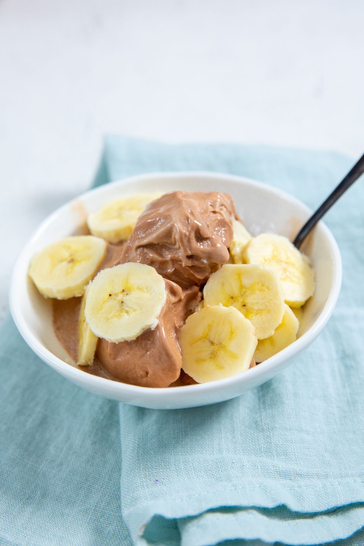 Chocolate Banana Protein Ice Cream in a white bowl with banana slices on top and a spoon in it with a blue napkin next to it