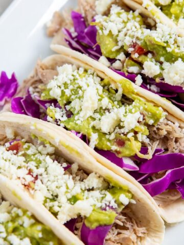 Low carb tacos filled with shredded chicken, purple cabbage, guacamole, and crumbled cheese on a white rectangular plate.