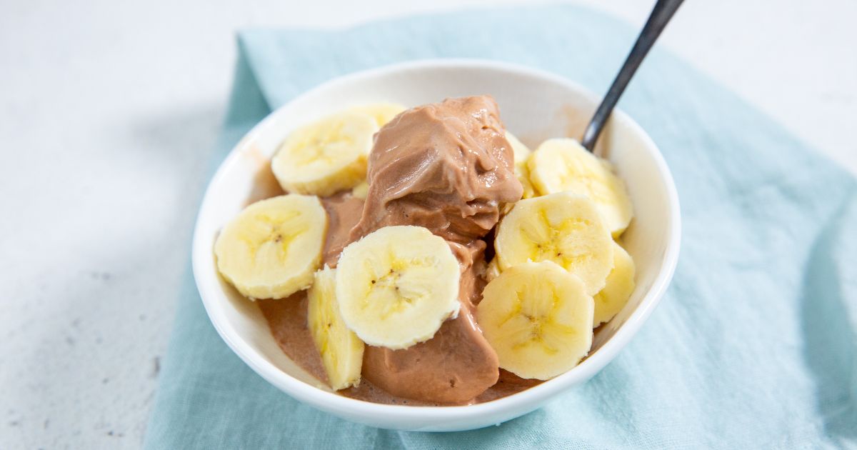 Chocolate Banana Protein Ice Cream in a white bowl with banana slices on top and a spoon in it with a blue napkin next to it