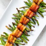 Eight bundles of green beans wrapped in crispy bacon are neatly arranged on a white rectangular plate.