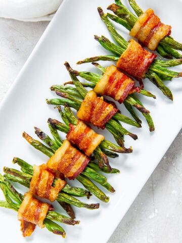 Eight bundles of green beans wrapped in crispy bacon are neatly arranged on a white rectangular plate.