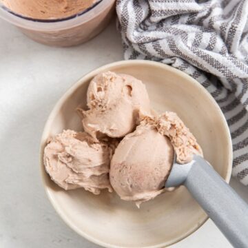 strawberry banana ice cream in a tan bowl with an ice cream scoop and a grey and white napkin